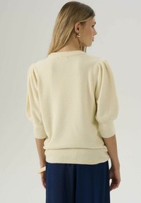 Un pull couleur crème avec un tissu texturé, des manches bouffantes et des poignets côtelés. Porté sur un pantalon bleu foncé, vu de dos.