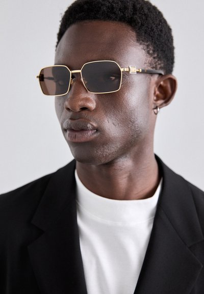 Gold-framed hexagonal sunglasses with brown tinted lenses, black temple accents, worn with a black blazer and white t-shirt.