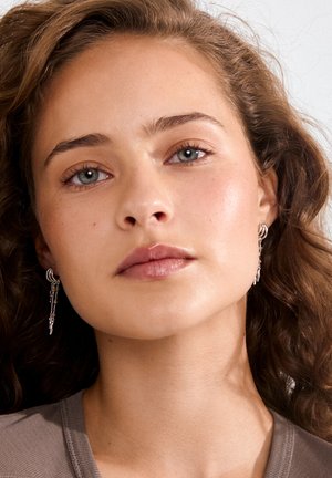 Mladá žena s kučeravými hnedými vlasmi, svetlomodrými očami, nosiace strieborné visiace náušnice a sivý top, tvárou k fotoaparátu na jednofarebnom pozadí.