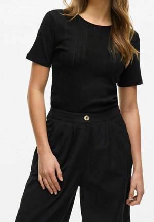 Kobieta w dopasowanym czarnym topie z krótkim rękawem i czarnych spodniach z wysokim stanem zapinanych na jeden guzik, stojąca na jednolitym tle.