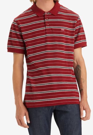 Man wearing a red, white, and black horizontal striped polo shirt with short sleeves and dark blue jeans.