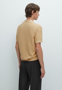 Polo a maniche corte, di un tan chiaro, con tessuto testurizzato e colletto a coste, abbinato a pantaloni scuri, visto da dietro.