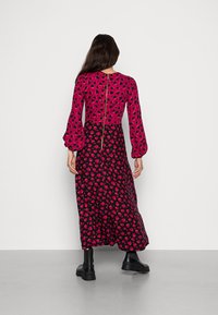 Lång klänning med en livlig rosa överdel med hjärtmönster, svart kjol med röda hjärtan, puffärm och synlig bakdragkedja. Svarta skor bärs.