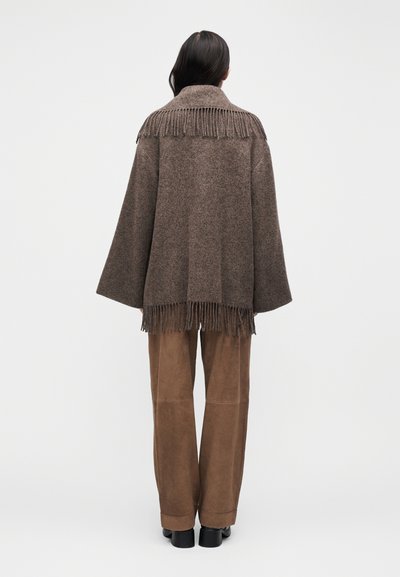 Barna gyapjú keverékből készült poncsó rojtos részletekkel a gallérnál és az alján. Laza szabás, széles ujjú. Világosbarna velúr nadrág és fekete cipő fölött viselve.