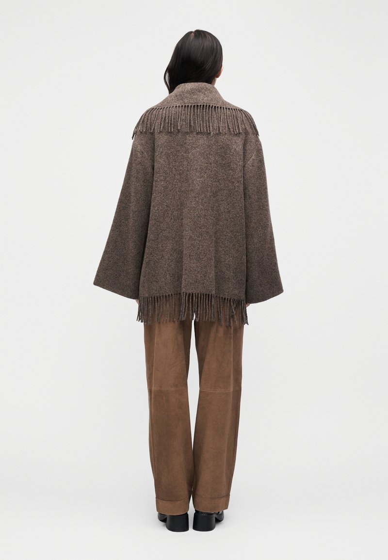Brun poncho i uldblanding med frynser ved kraven og kanten. Løs pasform, vide ærmer. Bær over tanfarvede ruskindsbukser og sorte sko.