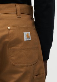 Τσέπη καφέ παντελονιού Carhartt με ορατό λογότυπο της μάρκας σε άτομο που φοράει μαύρο μακρυμάνικο πουκάμισο, κοντινή λήψη στην περιοχή της λεκάνης.