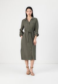 Vestido camisa verde oliva com botões na frente, cintura cinto, mangas dobradas e fendas laterais. Sandálias de tiras bege.