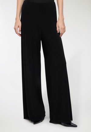 Pantaloni neri a gamba larga in morbido tessuto con vita elastica. Texture liscia e design a lunghezza completa, indossati con tacchi neri.
