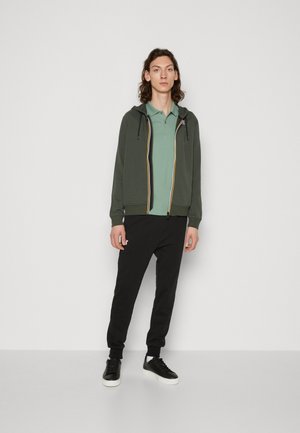 Hooded jack in olijfgroen met gele accenten, gecombineerd met een lichtgroen polo en zwarte joggingbroeken. Zwarte sneakers maken de outfit compleet.