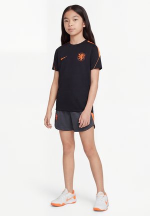 Joven niña de pie vestida con equipación de fútbol negra y naranja con el logo de Nike y zapatillas blancas con suelas naranjas.