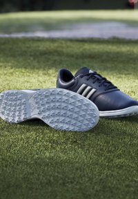 Ζεύγος άσπρα και μαύρα παπούτσια γκολφ Adidas σε πράσινο γρασίδι, ένα παπούτσι δείχνει την υφασμένη σόλα προς τα εμπρός.