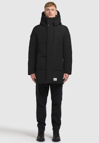 Svart vadderad jacka med hög krage, fyrkantig siluett och zipfickor i sidorna. Bärs över mörka byxor och stövlar. Slät, matt yta.