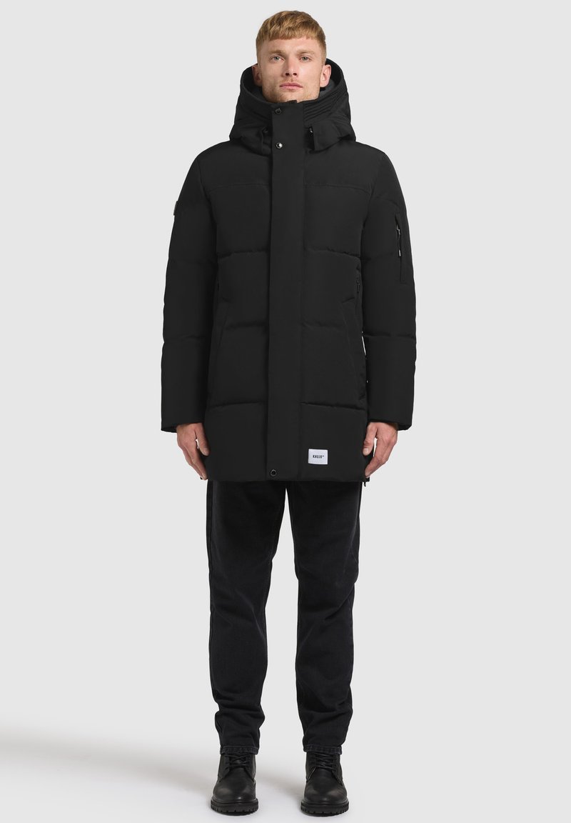 Svart vadderad jacka med hög krage, fyrkantig siluett och zipfickor i sidorna. Bärs över mörka byxor och stövlar. Slät, matt yta.