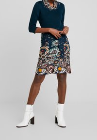 Stickad topp i djup teal med cowl-krage, kombinerad med en mönstrad marinblå kjol med blommönster och en knytning i midjan. Vita ankelboots.