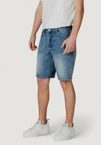 Pantalones cortos de mezclilla en azul claro con un diseño desgastado, estilo de cinco bolsillos y corte recto. Combinados con zapatillas blancas.