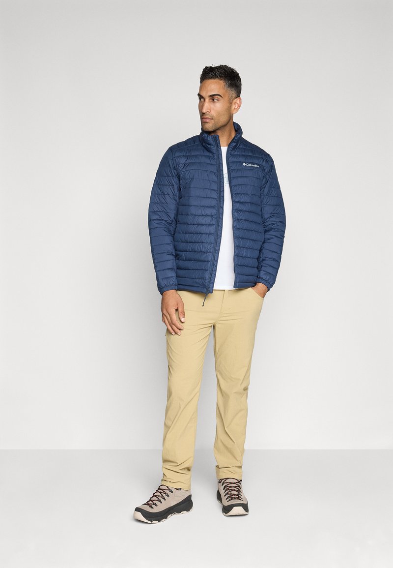 Veste matelassée bleu marine avec un col haut, assortie à une chemise blanche, un pantalon beige et des bottes de randonnée multicolores.