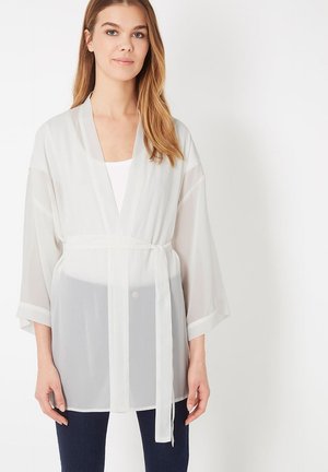 Mujer con una cárdigan estilo kimono blanca y translúcida, de mangas anchas y cinturón atado, sobre una blusa blanca y pantalones oscuros.