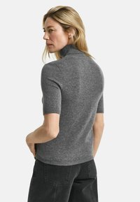 Frau mit blonden Haaren, die einen grauen kurzärmligen Rollkragenpullover und schwarze Jeans trägt, seitlich vor einem einfarbigen Hintergrund stehend.