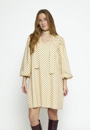 Woman wearing a cream-colored, long-sleeve, polka dot dress with a matching choker and dark knee-high boots, standing against a white background.