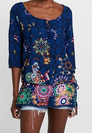 Frau trägt eine blaue Tunika mit bunten Mandala- und Blumenmustern, Dreiviertelärmeln und einem gebundenen Ausschnitt über ausgefransten Jeansshorts.