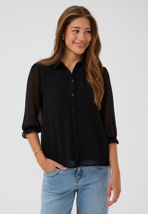 Woman with long hair wearing a sheer black button-up blouse with three-quarter sleeves and light blue jeans, smiling and looking sideways.