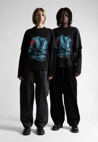 Melni kokvilnas T-krekli ar zilu māksliniecisku apdruku. Brīvi pieguļoši melni džinsi un melnas kurpes, kā arī minimālisma fons.