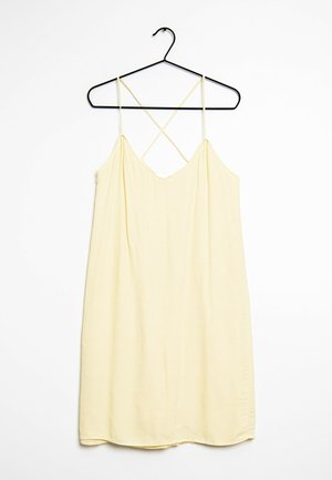 Żółta sukienka na ramiączkach z cienkimi, krzyżującymi się paskami, wykonana z miękkiego materiału, o delikatnym kroju w kształcie litery A i gładkiej fakturze. Wisi na czarnym wieszaku.