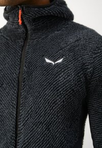 Fleece bunda s textúrovaným, tmavošedým exteriérom, vysokým golierom a čiernym zipsom. Má biele logo na hrudi a červené ťahátko zipsu.