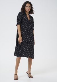 Zwarte jurk met stippen, met een relaxte pasvorm, korte pofmouwen en een V-hals. Glad materiaal met een rok tot op de knie.