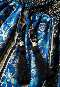 Un tissu à motifs bleus présente des designs floraux en noir et or. Il est agrémenté de franges noires avec des accents dorés nouées à la taille.