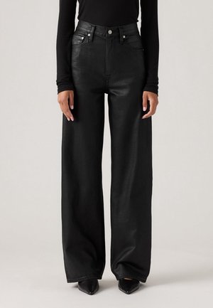 Personne portant un pantalon en cuir noir taille haute à jambes larges, associé à des chaussures noires pointues et un haut noir ajusté à manches longues.
