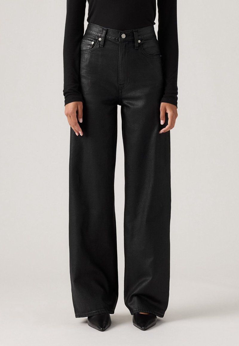 Personne portant un pantalon en cuir noir taille haute à jambes larges, associé à des chaussures noires pointues et un haut noir ajusté à manches longues.