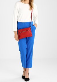 Czerwona, teksturowana torebka na ramię z detalem zamka błyskawicznego; noszona z białą bluzką z długim rękawem i niebieskimi, wysokim stanem spodniami. Czarne buty dopełniają strój.