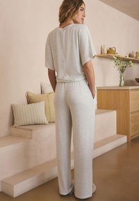Femme aux cheveux blonds ondulés portant une tenue décontractée gris clair, debout les mains dans les poches dans un espace de vie moderne aux tons beige.