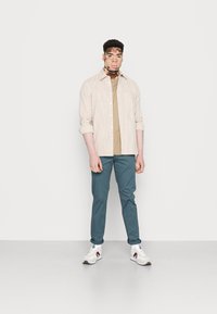 Chemise boutonnée beige rayée sur une chemise beige; pantalon bleu-vert et baskets blanches avec des accents rouges. Pose debout sur un fond uni.