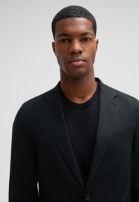 Junger Mann mit kurzem Haar, der einen schwarzen Blazer und ein schwarzes Hemd trägt und mit dem Gesicht gegen einen einfarbigen hellgrauen Hintergrund schaut.