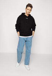 Felpa nera con cappuccio e tasca frontale, abbinata a jeans azzurri larghi e sneakers bianche. Logo semplice sul petto.