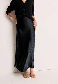 Fustă din satin negru cu un design fluid, textură netedă și o ușoară strălucire. Asortată cu o bluză neagră cu butoni și sandale cu toc nude.