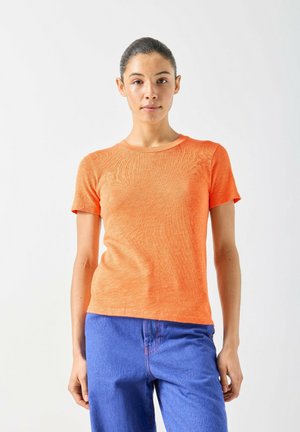 Femme aux cheveux tirés en arrière portant une chemise orange à manches courtes et un pantalon bleu, debout devant un fond clair uni.