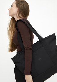 Svart axelväska i hållbart tyg med robusta remmar, med en enkel design och en lätt lyster. Logotypen är präglad på framsidan.