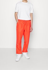 Man som bär klara orangea dragkedjebyxor, vita sneakers och en helt vanlig vit t-shirt med kort ärm, står mot en vit vägg.