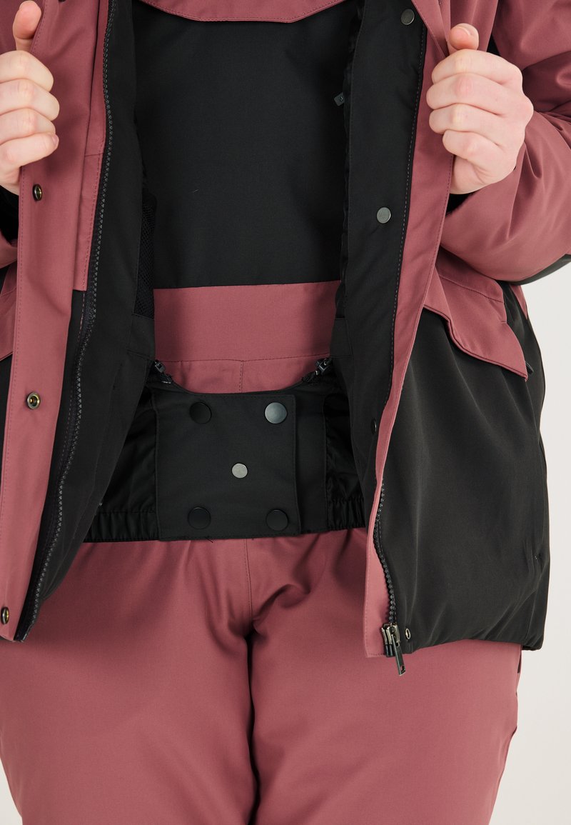 Utomhusjacka med rosa och svart färgschema, med tryckknappsstängningar och elastisk midja, som framhäver slitstarkt, texturerat tyg.