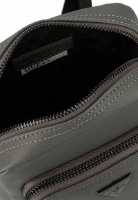 Grau synthetisches Materialtasche mit einem Reißverschluss. Das Innere hat ein dunkles Muster und ein sichtbares Markenetikett. Genähte Akzente verbessern die Struktur.