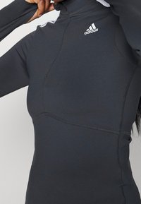 Persona che regola il colletto di una maglia sportiva nera a maniche lunghe Adidas ben aderente, con cuciture visibili e logo sul petto.