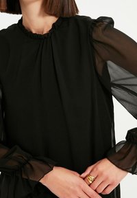 Schwarze Bluse mit voluminösem Ausschnitt und transparenten langen Ärmeln. Der Stoff ist leicht und hat eine glatte Textur, mit gerafften Manschetten an den Handgelenken.