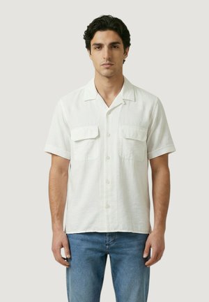 Homme portant une chemise blanche à manches courtes avec deux poches poitrine, associée à un jean bleu, debout devant un fond uni.
