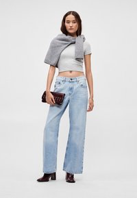 Φαρδιά τζιν παντελόνια σε ανοιχτό μπλε με ψηλή μέση, συνδυασμένα με γκρι cropped τοπ και γκρι πλεκτό κασκόλ που είναι περασμένο στους ώμους. Κρατάει ένα σκοτεινό μπορντό τσάντα χειρός.