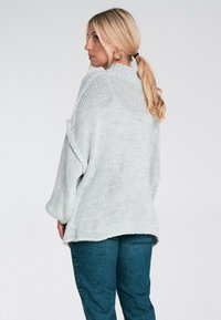 Jasnoszara, oversize'owa, dzianinowa sweter z długimi rękawami i opadłymi ramionami, zestawiona z niebieskimi dżinsami. Charakteryzuje się luźnym, teksturowanym wzorem.