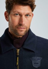 Námornícka zipová bunda s vysokým límcem, vlněnou texturou, vyšitým logem a mosazným zipem. Tmavě vínová vnitřní podšívka viditelná.