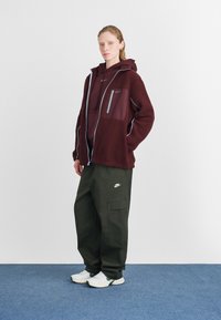 Burgunderfleecejakke med grå glidelås lommer over en mørk burgunder hettegenser, kombinert med olivengrønne cargo-bukser og hvite joggesko.